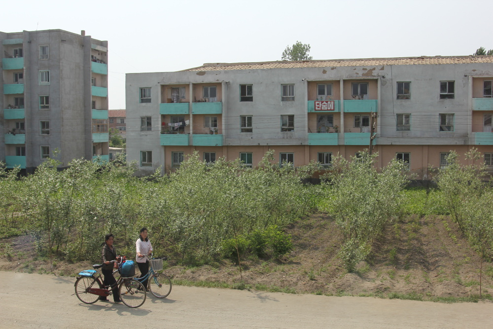 Zwei Nordkoreaner schieben ihr Fahrrad über eine staubige Piste, hinten Wohngebäude